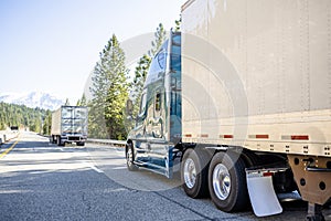 Convoy of the long haulers big rigs industrial semi trucks with semi trailers driving on the straight highway road with forest on