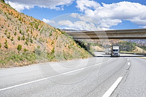 Convoy of two big rigs semi trucks with loaded semi trailers driving on the turning mountain highway road going under the bridge