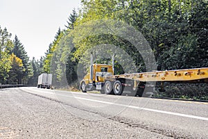 Convoy of two different big rig semi trucks with flat bed and dry van semi trailers driving on the winding narrow road with forest