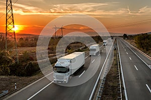 Convoy of white lorry trucks on a highway at sunset