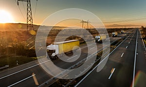 Convoy of white transportation trucks with yellow trailers in line on a country highway at sunset