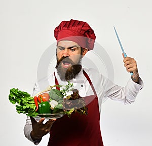 Cook with angry face in burgundy uniform cuts salad ingredients.