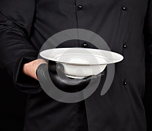 Cook in black uniform and black latex gloves holds in his hand a round empty white plate