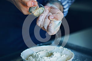 A cook in a blue apron holds a salt shaker in hands, and pours the seasoning into the flour in a dough bowl. The process of