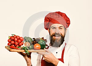 Cook with cheerful face in burgundy uniform holds salad ingredients. Chef holds board with fresh vegetables. Cooking and