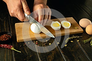 A cook cuts a boiled egg with a knife before preparing an eggs dish. Low key calorie diet concept on kitchen table. Place for