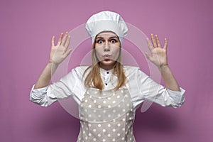 Cook girl in apron raises and shows hands on a pink background, scared woman housewife in kitchen clothes