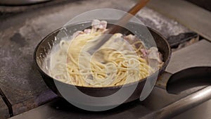 Cook holding stirring spaghetti with cheese sauce and chopped bacon frying in hot pan