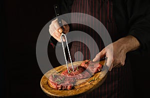 The cook holds fork in his hand and a cutting board with beef steaks before barbecue. Black space for restaurant recipe or hotel