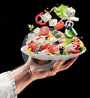 Cook holds white plate of greek salad on black background. Ingredients of salad falling down om the plate. Tasty food background