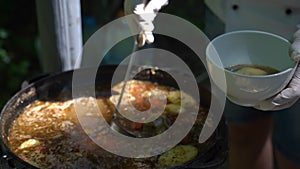 The cook pours meat soup in a cast-iron cauldron on plates. Slow motion