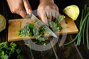 The cook is preparing a salad on the kitchen table. Close-up of a chef hands using a knife to chop fresh green parsley