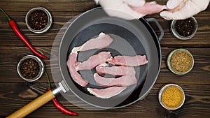 The cook puts the meat on the pan. Sliced pork in a cast iron skillet. A metal frying pan on a wooden table surrounded by spices