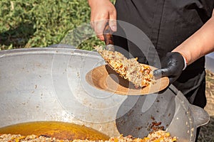 Cook puts pilaf with meat and carrots on a disposable paper plate against the backdrop of a large cast iron cauldron.