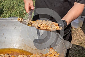 Cook puts pilaf with meat and carrots on a disposable paper plate against the backdrop of a large cast iron cauldron.