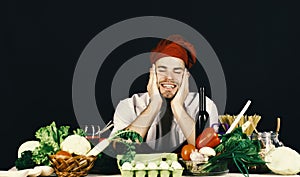 Cook sits in kitchen near table with vegetables and tools. Cuisine and professional cooking. Man in burgundy hat and