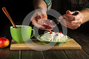 The cook with a spoon add sweet jam to milk yogurt and cheese on a kitchen black table while preparing a diet meal