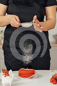 The cook sprinkles flour on a red cake through a sieve