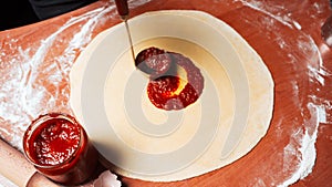 The cook takes tomato sauce from glass jar into ladle and places it on raw dough