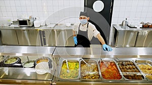 A cook or waiter, in protective gloves and mask, stands behind display case with different dishes, in self-service