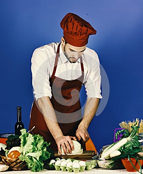 Cook works in kitchen near table with vegetables and tools. Chef with busy face chops cabbage with knife on blue