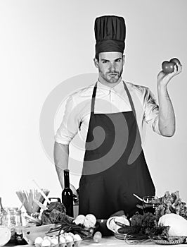 Cook works in kitchen near table with vegetables and tools. Cuisine and cooking concept. Man in cook hat and apron