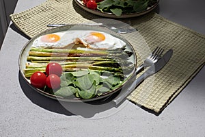 Cooked asparagus with fresh spinach leaves, tomatoes and scrambled eggs on a green plate on a table with a kitchen towel