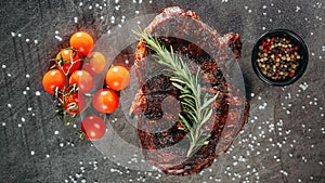 Cooked beef steak on a dark metal cutting board, next to a sprig of aromatic rosemary, tomatoes and a black pot of peppercorn mix
