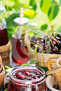 Cooked homemade cherry jam in glass jar on white wooden table outdoors, fresh cherry jam close up