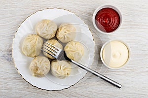 Cooked khinkali, fork in plate, bowls with ketchup and mayonnaise on wooden table. Top view