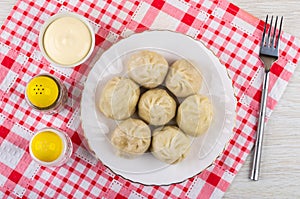 Cooked khinkali in plate, bowl with mayonnaise, shakers with salt and pepper, fork on checkered napkin on wooden table. Top