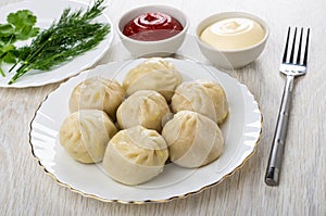 Cooked khinkali in plate, bowls with ketchup and mayonnaise, greens in plate, fork on wooden table