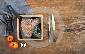 Cooked meat t-bone steak with garlic cloves, tomatoes, rosemary, pepper and salt in small cooking pan over rustic wooden