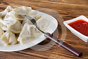 Cooked Russian dumplings in white plate with fork next to red sauce on natural wooden pine boards. Photo