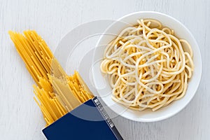 Cooked spaghetti in a white ceramic bowl next to uncooked spaghetti in a blue box on white painted wood