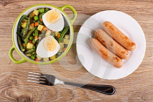 Vegetable mix, boiled egg in bowl, knife, salt shaker, pepper shaker, fried sausages in plate, fork on table. Top view