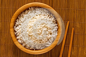 Cooked white rice in a dark wood bowl next to chopsticks isolated on bamboo matt from above