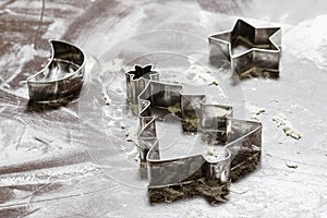 The cookie cutters in the shape of `christmas tree`, `star` and `half moon` together with flour lie on the wooden table