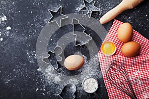 Cookies cutters, flour, rolling pin, eggs and whisk on kitchen table top view. Christmas baking background.