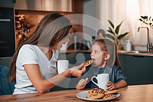 With cookies and drink. Young mother with her daughter is together at apartment