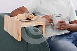 Cookies on sofa armrest wooden table. Man holding cup of drink at home, closeup