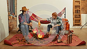 Cooking chicken over fake fire. Two men imagining, recreating camping activity indoors with necessary equipment. Concept