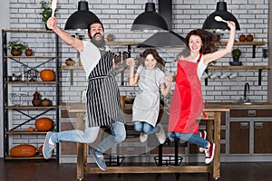 Cooking with child might be fun. Having fun in kitchen. Family mom dad and little daughter wear aprons jump in kitchen