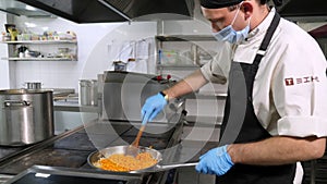 Cooking. close-up. the cook, in protective mask and gloves, stirring the frying onions and carrots in frying pan. health