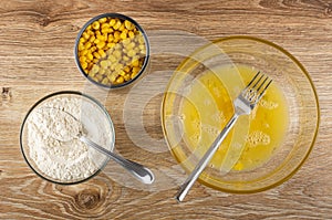 Cooking corn pancakes. Jar of corn, spoon in bowl with flour, fork in bowl with scrambled eggs on table. Top view