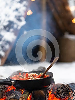 Cooking food in pan on fire near survival shelter in winter forest.