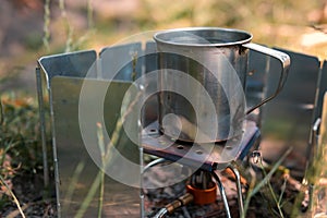 Cooking on the go. Tourist equipment, gas burner, wind protection, metal cup with water, green grass. Selective focus. Summer day