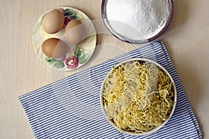 Cooking homemade noodles, flat lay. On wooden surface ingredients are wheat flour and eggs, and finished product