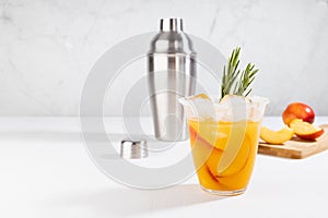 Cooking of refreshing juicy peach cocktail with ice, rosemary, sugar rim in misted glass, ingredients, silver shaker on white wood