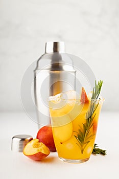 Cooking of refreshing juicy peach cocktail with ice, rosemary, sugar rim in misted glass, ingredients, silver shaker on white wood